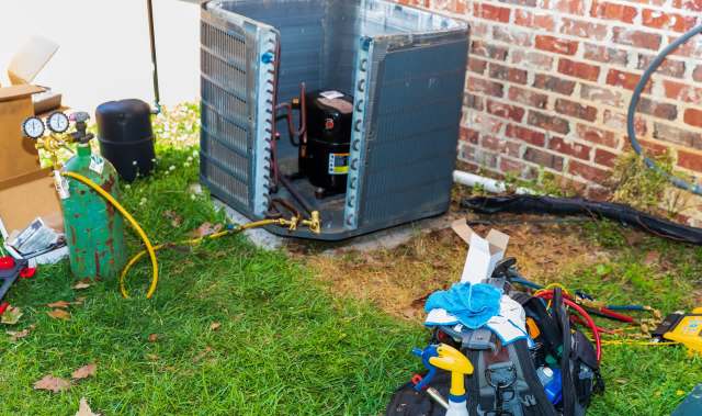 An outdoor scene featuring an open air conditioning unit, tools, and a green gas cylinder on grass.