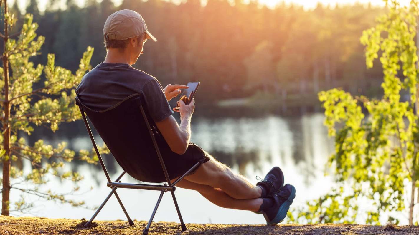 A person sitting in a chair by a serene lake, using a smartphone, surrounded by trees at sunset.