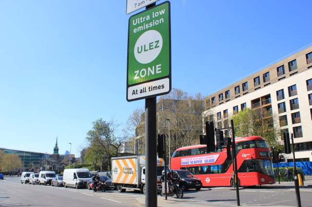 A street scene featuring a ULEZ sign, buses, and parked vehicles under a clear blue sky.