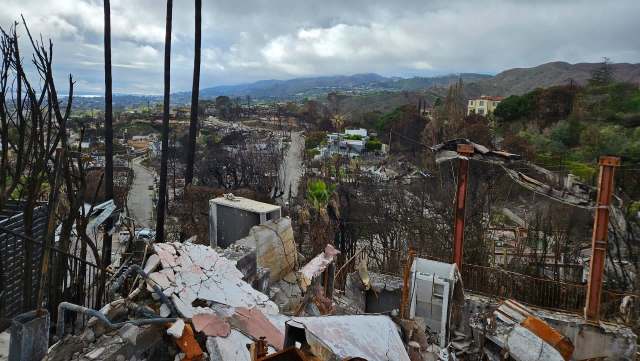 A devastated landscape shows burnt structures, barren trees, and a road winding through a fire-damaged neighborhood.