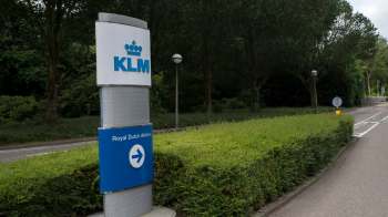 KLM sign indicating Royal Dutch Airlines, surrounded by greenery and a road leading away from it.