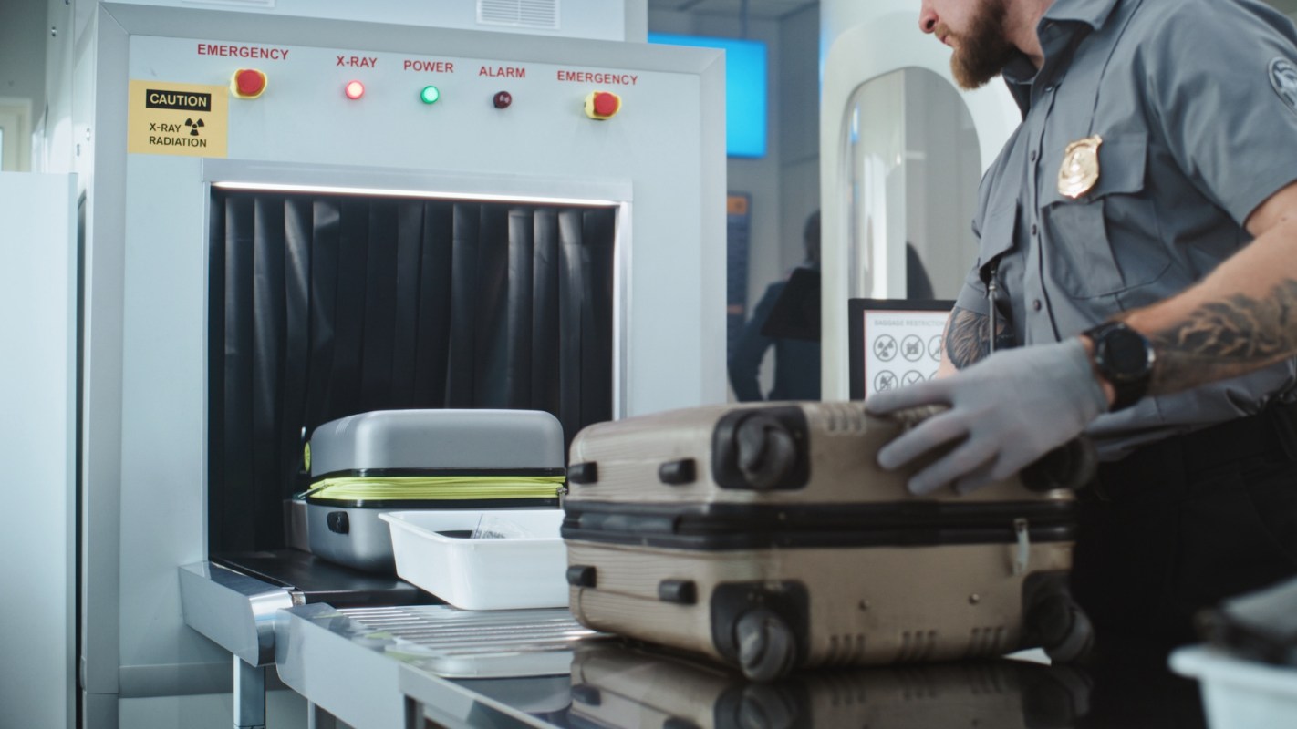 A security officer inspects baggage at an airport screening station with X-ray equipment in the background.