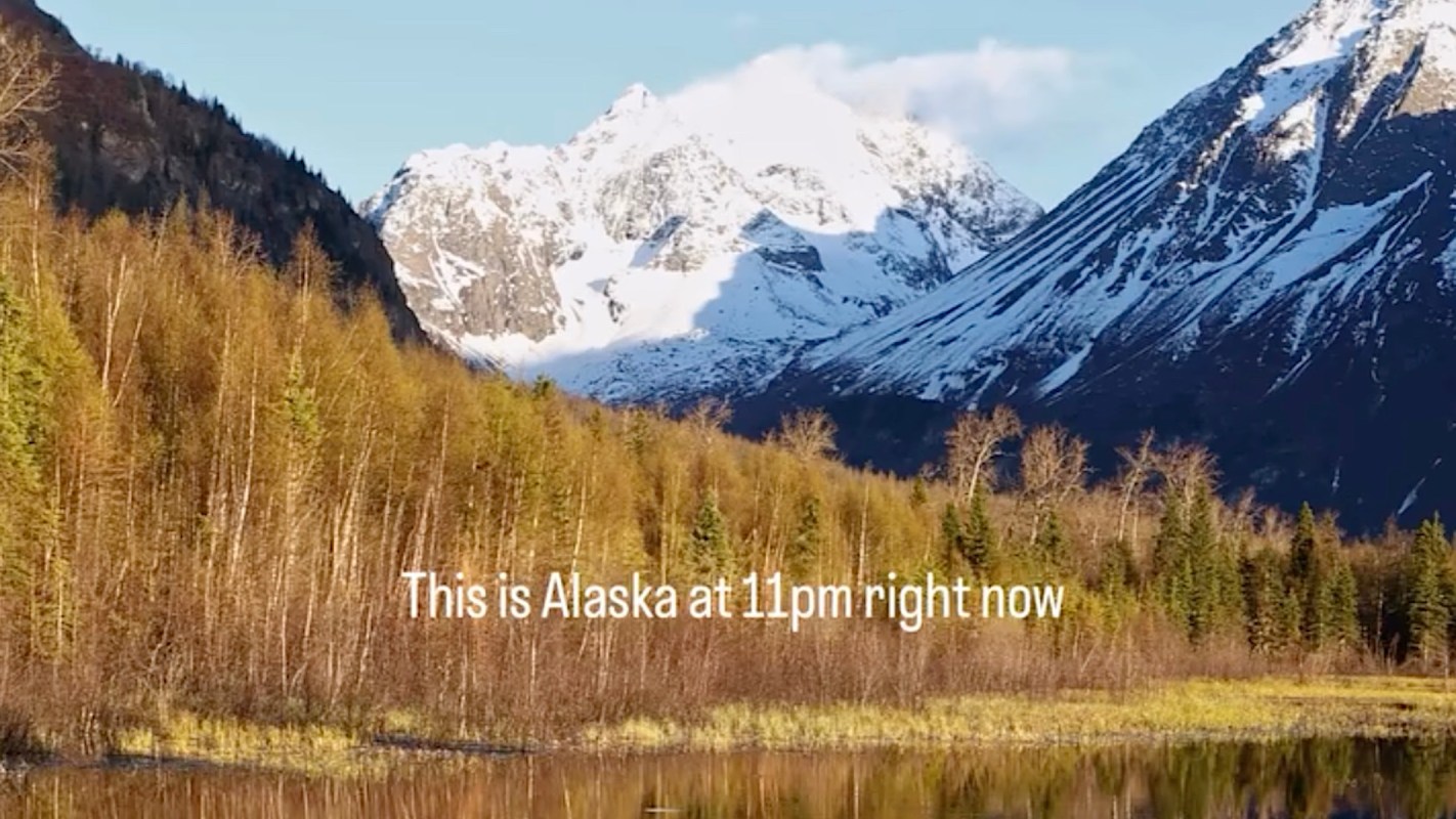 A serene landscape of snow-capped mountains reflected in a calm lake surrounded by evergreen trees at dusk.