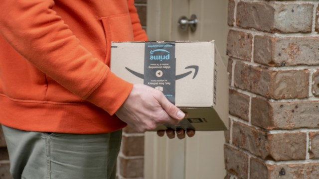 A person wearing an orange hoodie holds an Amazon package outside a doorway with a brick wall background.