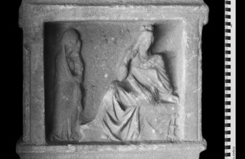 A stone relief depicting two figures, one seated and another standing, in a classical style.