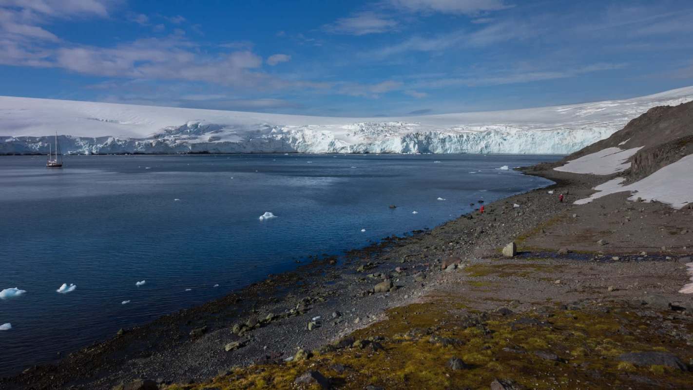 A serene coastal landscape in Antarctica featuring a glacier, a calm sea, and a small sailing boat in the distance.