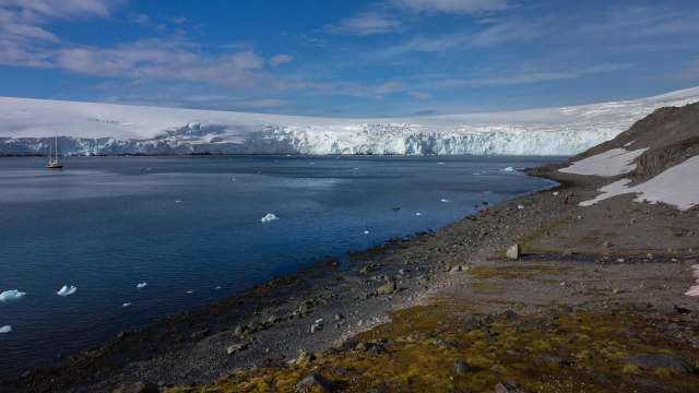 A serene coastal landscape in Antarctica featuring a glacier, a calm sea, and a small sailing boat in the distance.