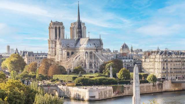 A view of the Notre-Dame Cathedral in Paris surrounded by trees and buildings along the Seine River under a clear blue sky.