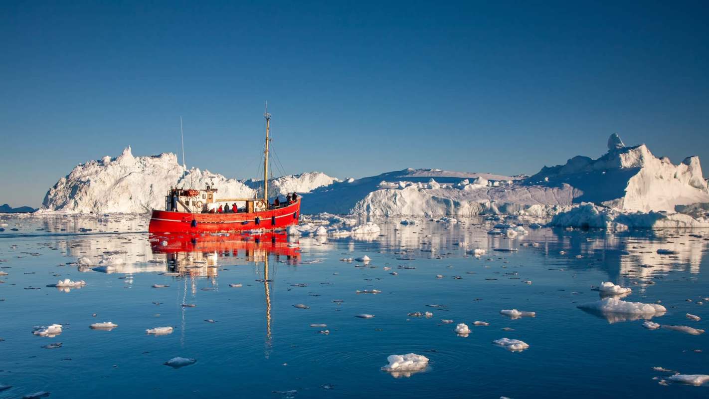 A red boat navigates icy waters surrounded by large icebergs under a clear blue sky.