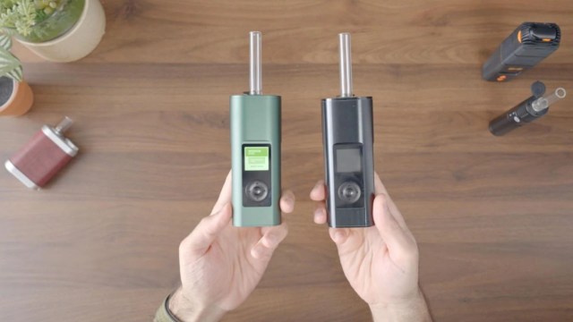 Two vaporizers, one green and one black, held in hands over a wooden table with a plant and other devices nearby.