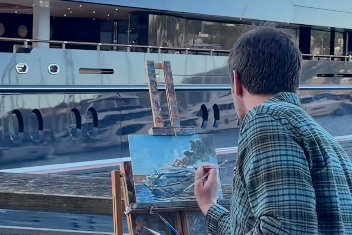An artist paints a yacht on an easel beside a marina, with a luxurious boat reflected in the water.