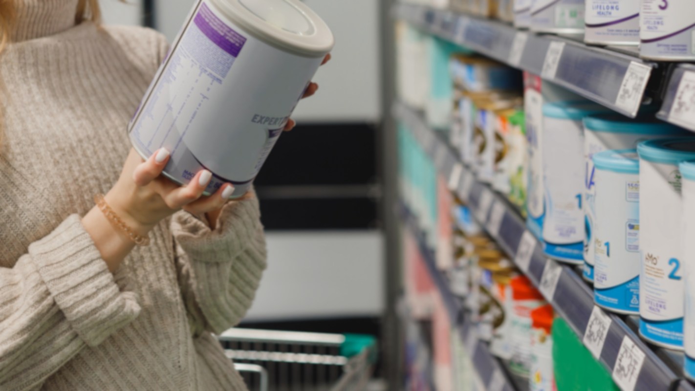 A person in a beige sweater examines a container while shopping in a store aisle filled with various products.