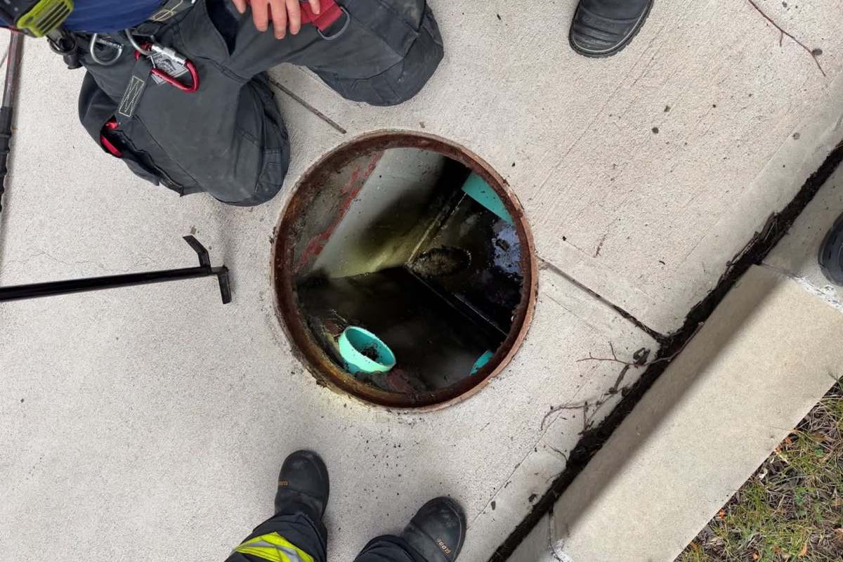 Two individuals stand beside an open manhole on a concrete sidewalk, with tools nearby and a view into the dark chamber below.