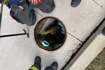 Two individuals stand beside an open manhole on a concrete sidewalk, with tools nearby and a view into the dark chamber below.