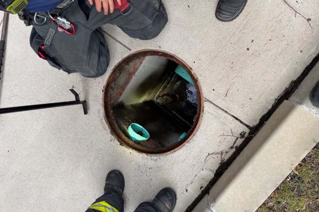 Two individuals stand beside an open manhole on a concrete sidewalk, with tools nearby and a view into the dark chamber below.