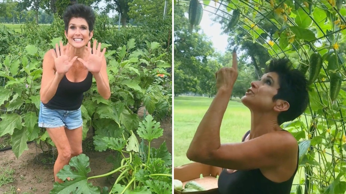 A woman in a garden enthusiastically gestures with her hands, surrounded by lush green plants and vegetables.