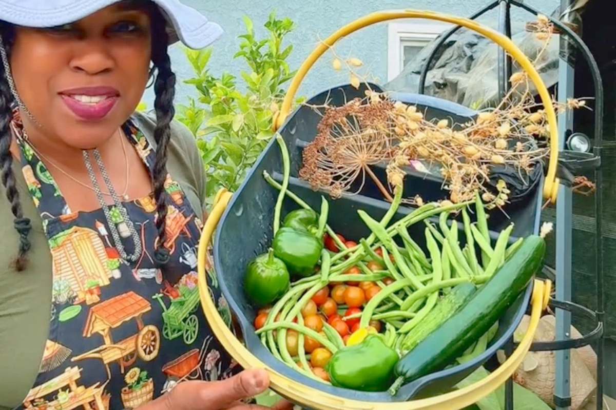 A woman in a garden proudly holds a basket filled with colorful vegetables, including peppers, tomatoes, and cucumbers.