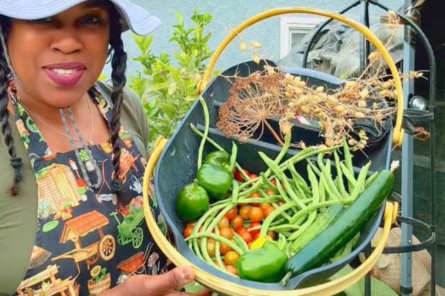 A woman in a garden proudly holds a basket filled with colorful vegetables, including peppers, tomatoes, and cucumbers.