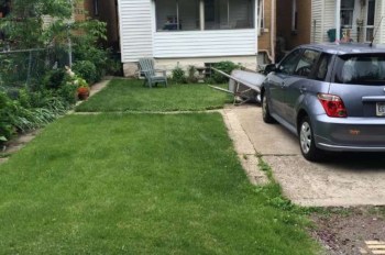 A car parked on a driveway next to a grassy area and a wooden bench beside a house.