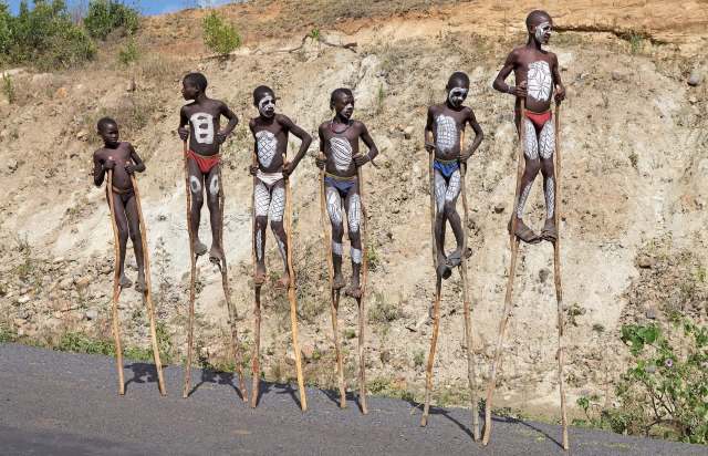 Six children, painted with traditional designs, stand on stilts in front of a dry, hilly landscape.