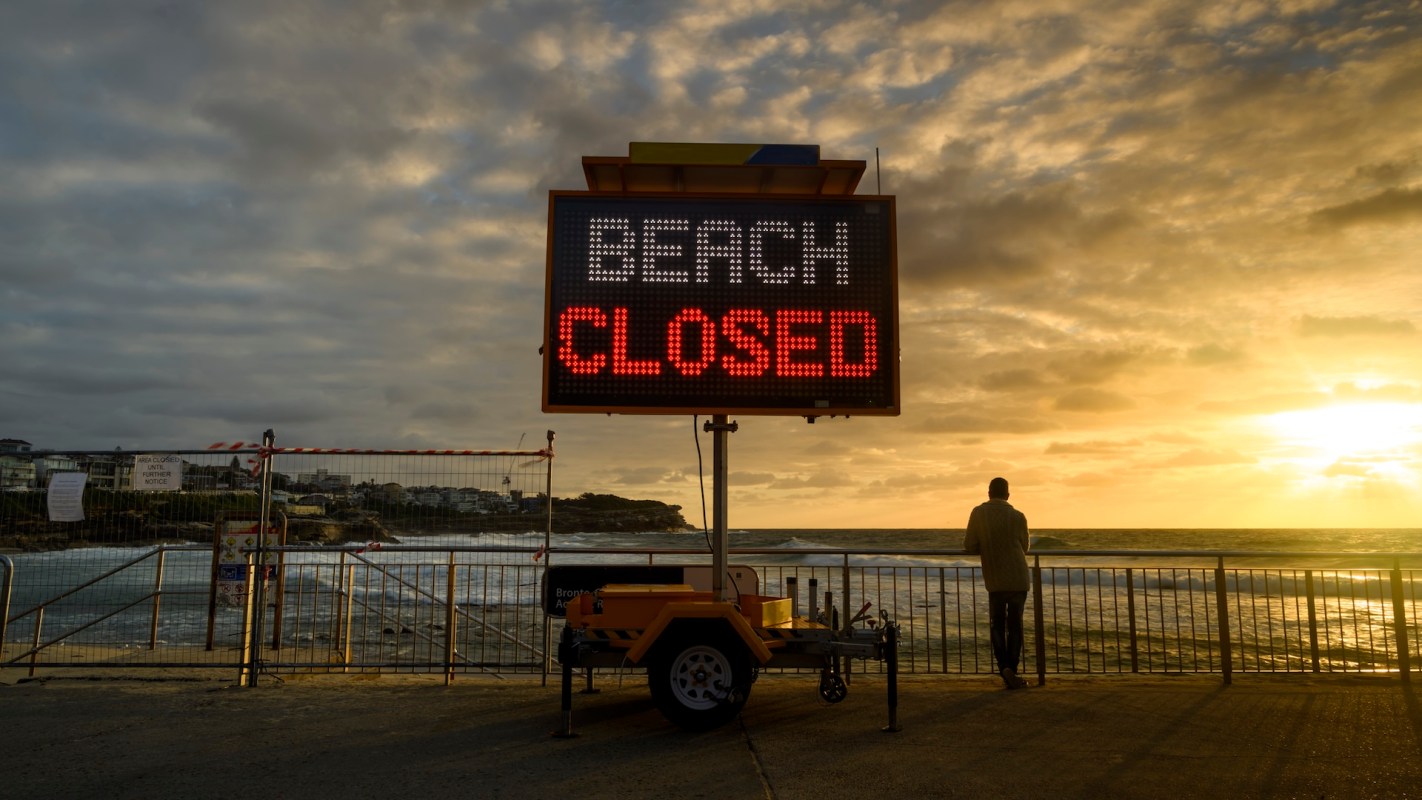 A man stands by a closed beach sign at sunset, with waves and clouds in the background.