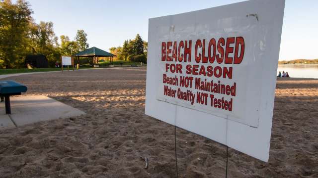 A sign warning that the beach is closed for the season, located in a sandy area near a lake.