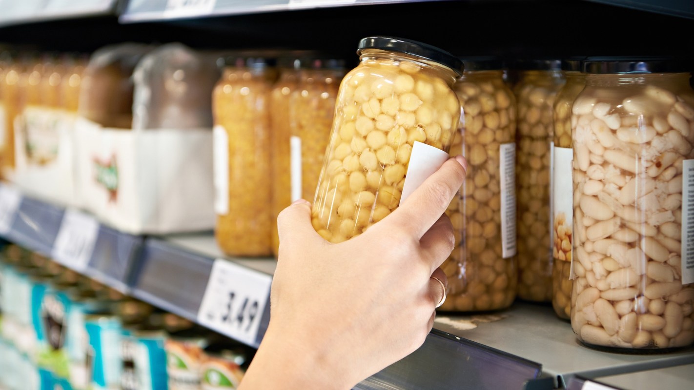 A hand reaches for a jar of chickpeas on a grocery store shelf filled with various canned goods.
