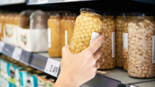 A hand reaches for a jar of chickpeas on a grocery store shelf filled with various canned goods.