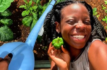 A smiling woman holds a green bell pepper in a sunlit garden with leafy plants and a blue watering can nearby.
