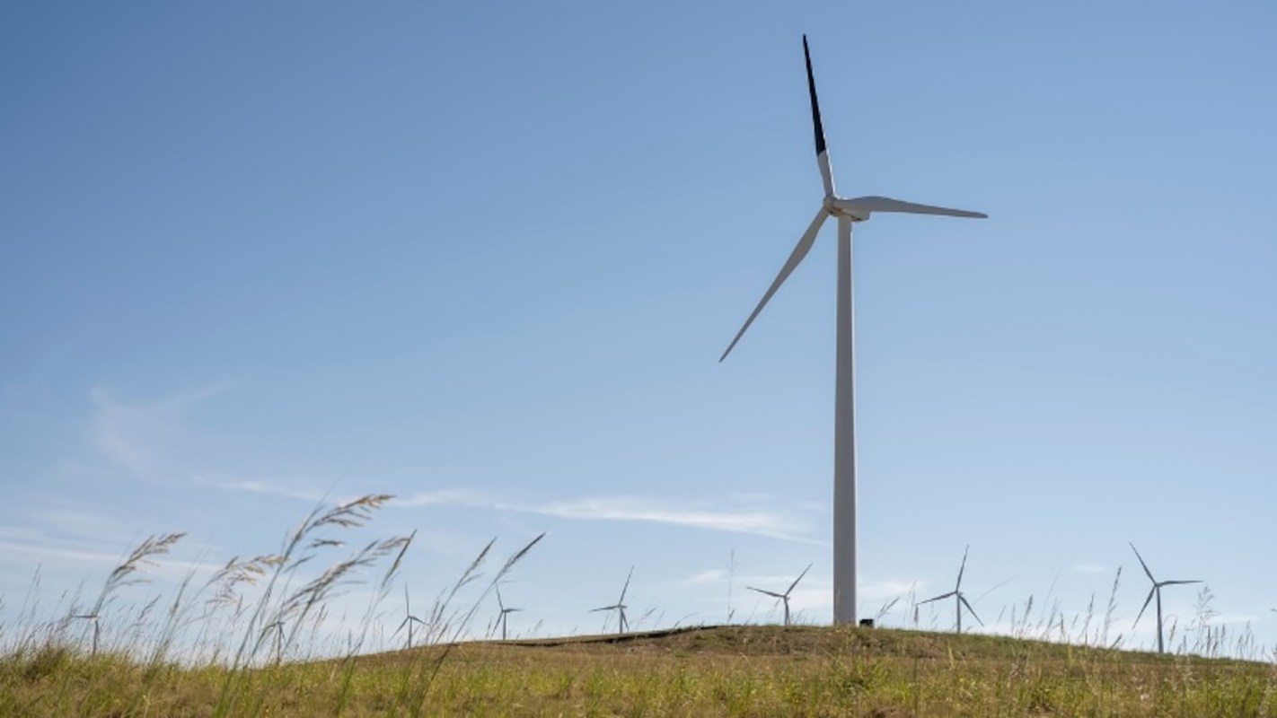 A wind turbine on a grassy hill against a blue sky, with one blade painted black and other turbines in the background.