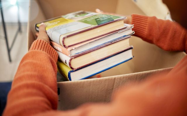 A person wearing an orange sweater holds a stack of books above an open cardboard box.