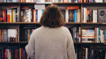 A person with shoulder-length hair stands in front of a bookshelf filled with various books.