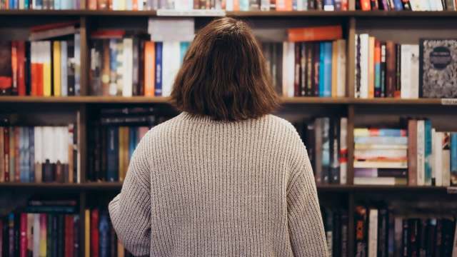 A person with shoulder-length hair stands in front of a bookshelf filled with various books.