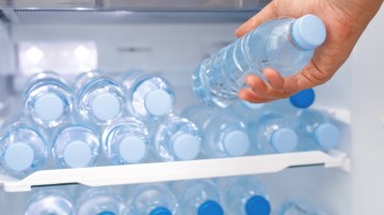 A hand reaches for a bottle of water inside a refrigerator filled with multiple similar bottles.