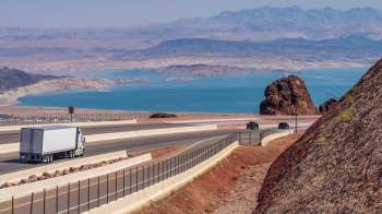 A truck and car drive along a winding highway with a blue lake and mountains in the background.