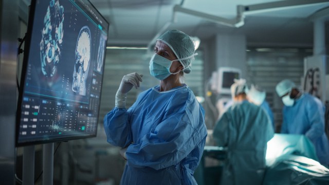 A surgeon in scrubs observes brain scans on a monitor in a surgical setting.