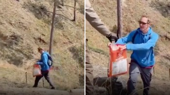 A person in a blue hoodie carries a large orange bag along a rocky, wooded trail.