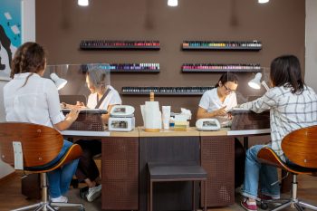Two nail technicians work on clients in a modern salon with a variety of nail polish displayed on shelves.