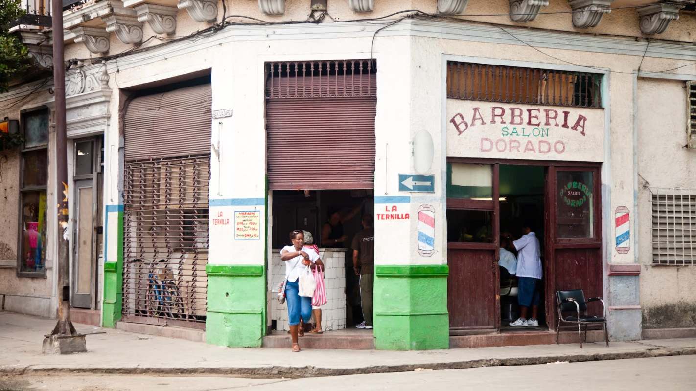 A woman exits a barber shop with a bag, while two people are inside getting a haircut.