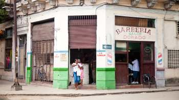 A woman exits a barber shop with a bag, while two people are inside getting a haircut.