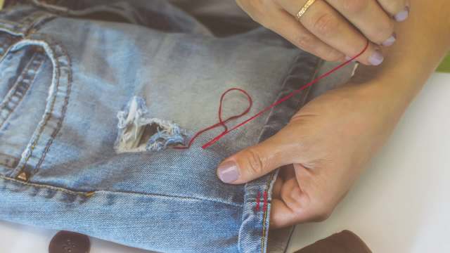 A person stitching a tear on blue jeans with red thread.