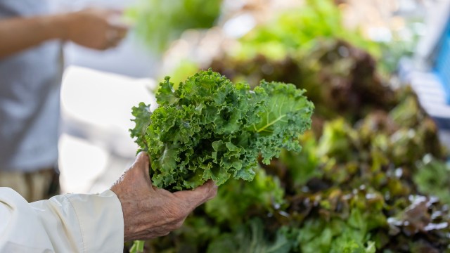 A person holds a bunch of fresh kale in front of a display of various leafy greens at a market.