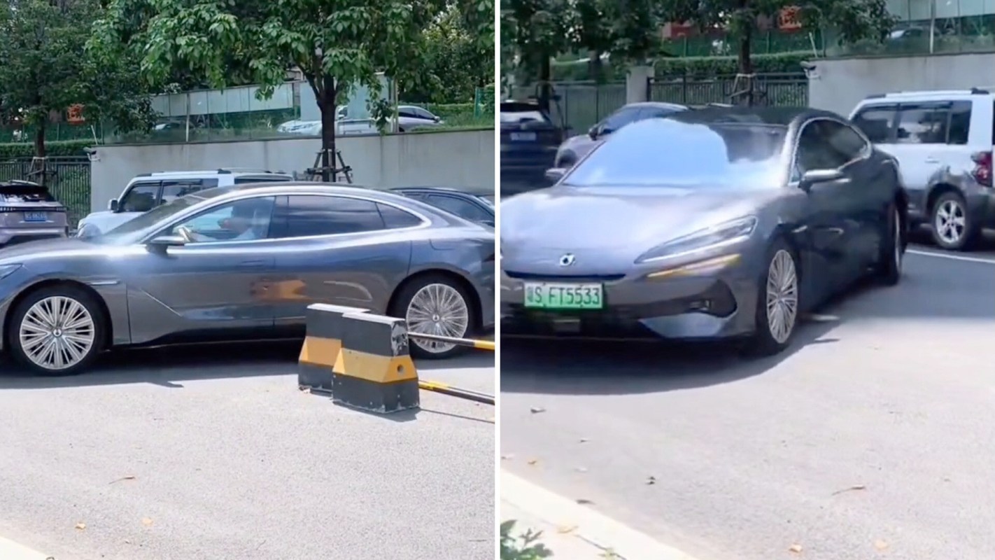 A sleek gray car navigates around a parking barrier in a busy outdoor parking lot.