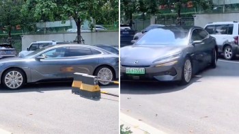A sleek gray car navigates around a parking barrier in a busy outdoor parking lot.