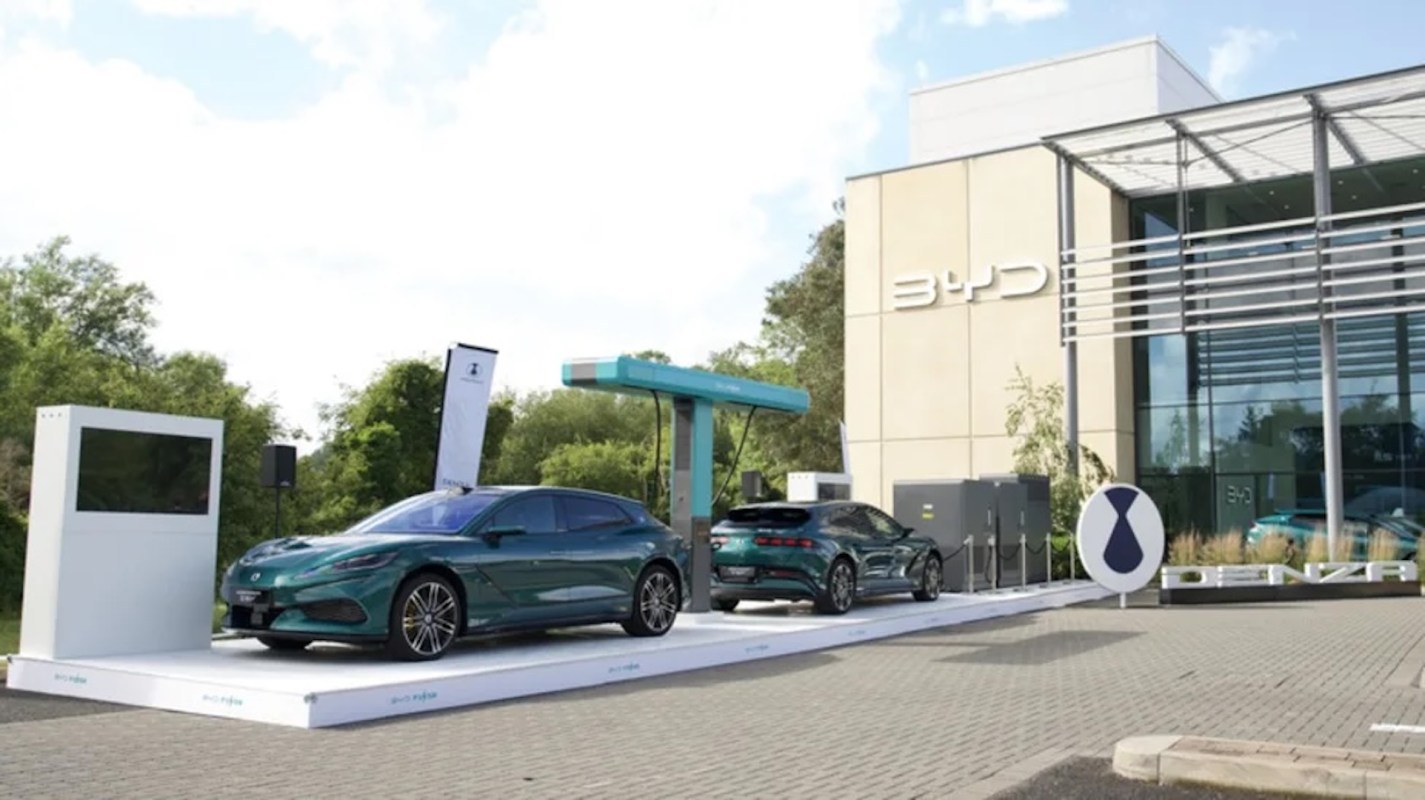 Two electric cars on display outside a BYD building, next to a charging station and promotional signage.