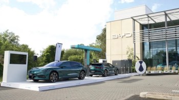 Two electric cars on display outside a BYD building, next to a charging station and promotional signage.