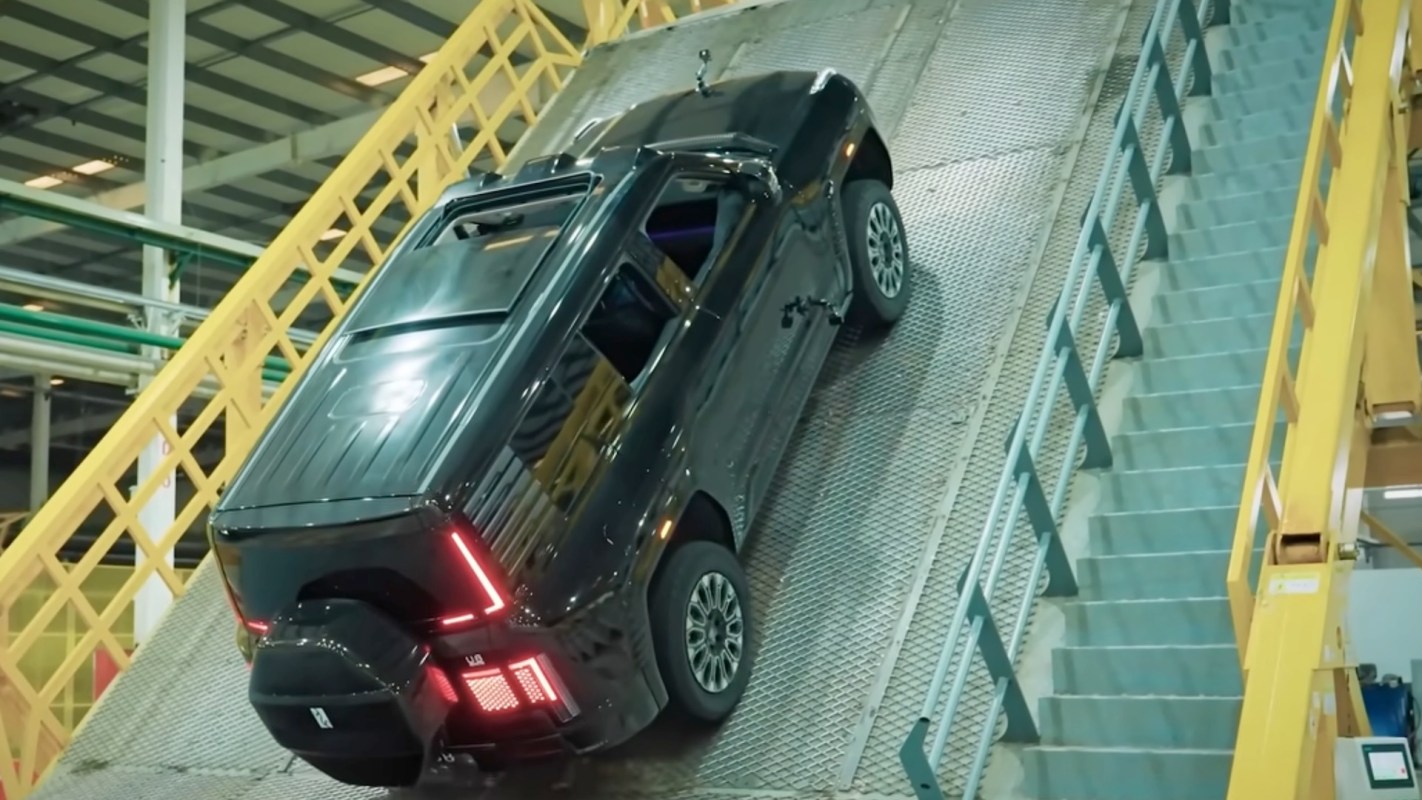 A black SUV is ascending a steep metal ramp in a factory setting.