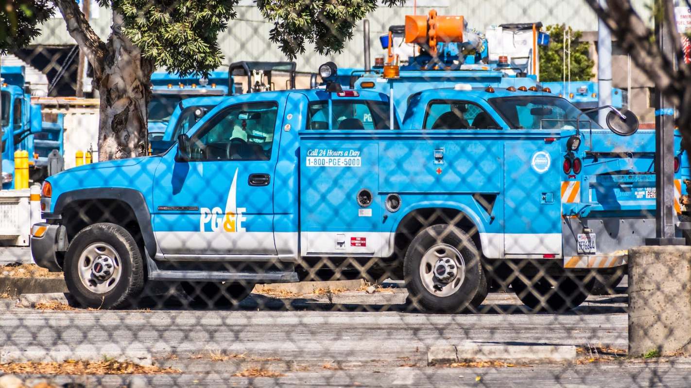 A blue utility truck marked with "PG&E" parked behind a chain-link fence among other vehicles.