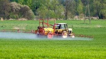 A yellow tractor spraying crops in a green field with trees in the background.