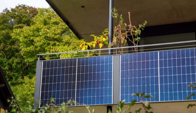 A balcony with solar panels and potted plants, set against a backdrop of lush green trees.